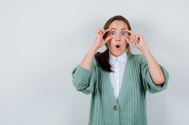 Mujer joven en blusa, chaqueta de punto abriendo los ojos con los dedos y mirando maravillado, vista frontal.