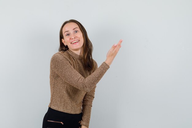Mujer joven en blusa dorada levantando la mano mientras muestra algo en el lado izquierdo y parece complacida, vista frontal. espacio para texto