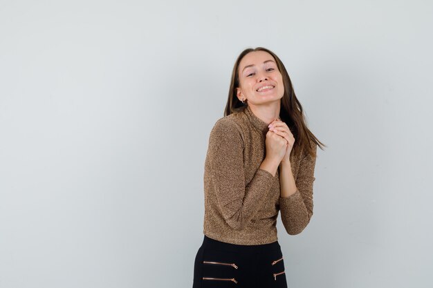 Mujer joven en blusa dorada sosteniendo las manos combinadas sobre su pecho y mirando alegre, vista frontal. espacio para texto