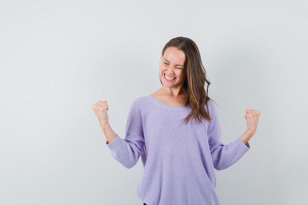 Mujer joven en blusa lila mostrando gesto ganador y mirando alegre