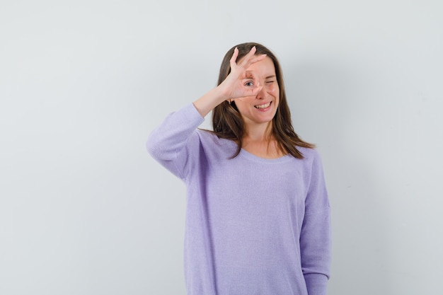 Mujer joven en blusa lila mostrando gesto ok sobre su ojo y mirando alegre