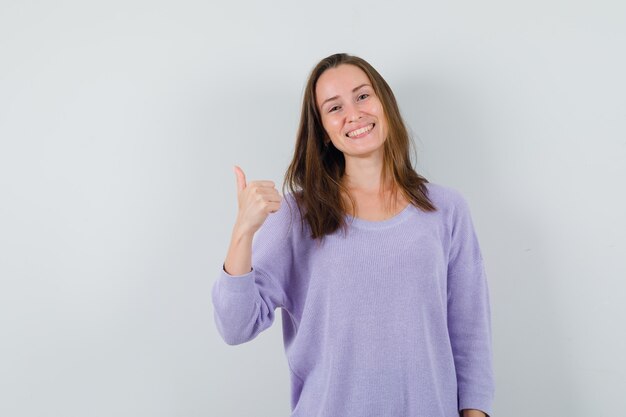 Mujer joven en blusa lila mostrando el pulgar hacia arriba y mirando alegre