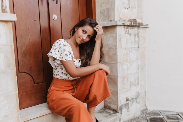 Mujer joven en blusa de manga corta y elegantes pantalones de cintura alta se sienta en la puerta