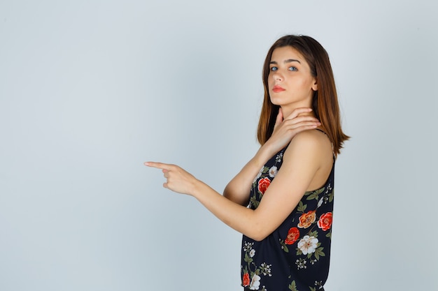 Mujer joven en blusa con la mano en el cuello, apuntando hacia adelante y mirando confiado.