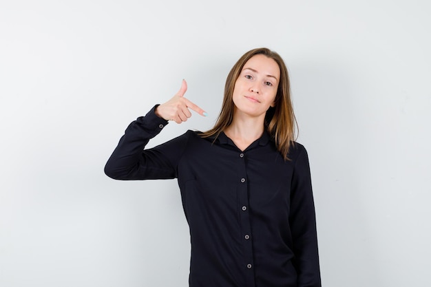 Mujer joven en blusa negra apuntando a sí misma con el dedo índice y mirando feliz, vista frontal.