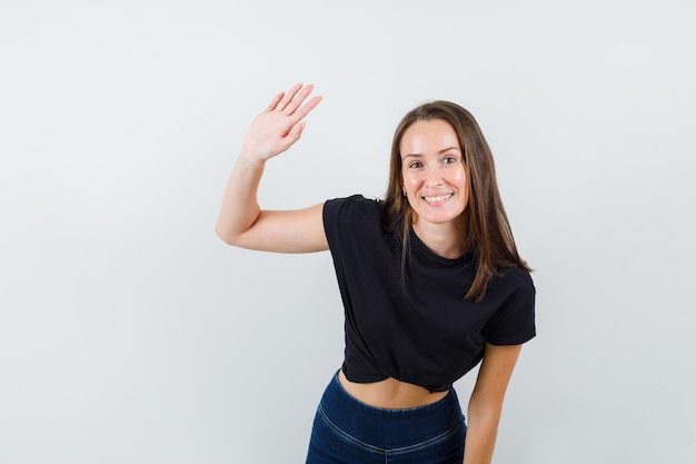 Mujer joven en blusa negra, pantalones agitando la mano para saludar y luciendo optimista