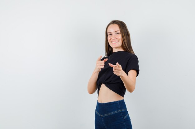 Mujer joven en blusa negra, pantalones apuntando a la cámara y mirando alegre