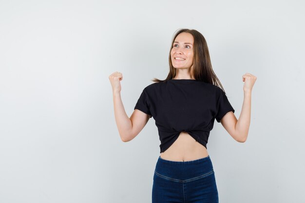 Mujer joven en blusa negra, pantalones mostrando gesto ganador y mirando feliz