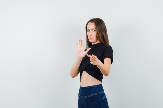 Mujer joven en blusa negra, pantalones mostrando la palma, apuntando a la cámara y mirando seguro
