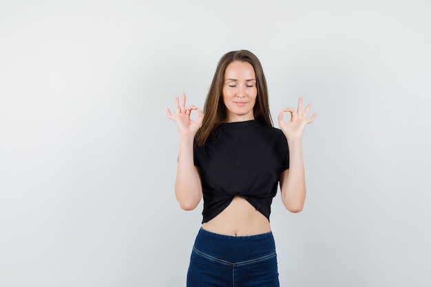 Mujer joven en blusa negra, pantalones mostrando signo de ok y buscando pacífica