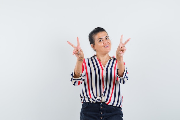 Mujer joven en blusa a rayas que muestra el signo de la paz con ambas manos y parece optimista