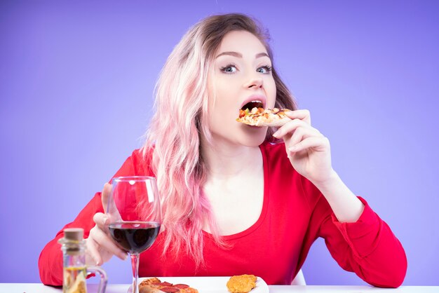 Mujer joven en blusa roja come pizza y sostiene una copa de vino tinto