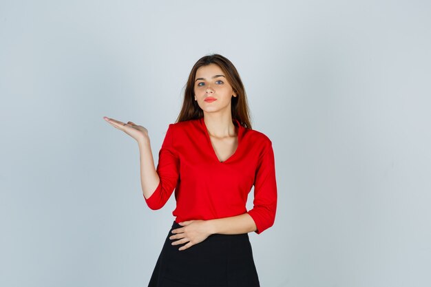 Mujer joven en blusa roja, falda negra extendiendo la palma hacia un lado y mirando seductor