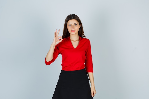 Mujer joven en blusa roja, falda negra mostrando signo ok y mirando feliz