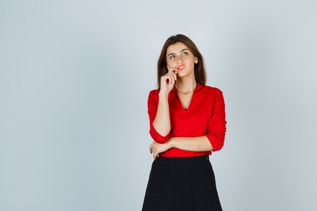 Mujer joven en blusa roja, falda negra de pie en pose de pensamiento