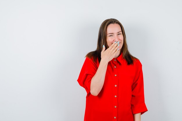 Mujer joven en blusa roja manteniendo la mano en la boca y mirando alegre, vista frontal.