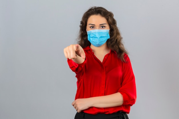 Mujer joven con blusa roja en máscara protectora médica apuntando a la cámara con el dedo índice mirando seriamente sobre fondo blanco aislado