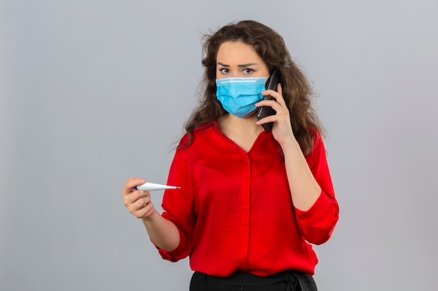 Mujer joven con blusa roja en máscara protectora médica hablando por teléfono móvil mirando preocupado sosteniendo termómetro digital sobre fondo blanco aislado