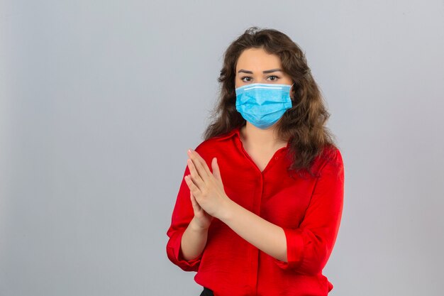 Mujer joven con blusa roja en máscara protectora médica mirando a la cámara frotándose las manos sobre fondo blanco aislado