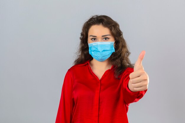 Mujer joven con blusa roja en máscara protectora médica mirando a la cámara con una sonrisa que muestra el pulgar hacia arriba sobre fondo blanco aislado