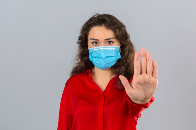 Mujer joven con blusa roja en máscara protectora médica de pie con la mano abierta haciendo señal de stop con gesto de defensa de expresión seria y segura sobre fondo blanco aislado