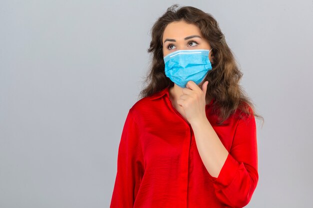 Mujer joven con blusa roja en máscara protectora médica de pie con la mano en la barbilla pensando tener dudas sobre fondo blanco aislado