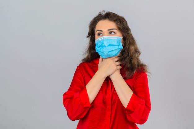 Mujer joven con blusa roja en máscara protectora médica que parece enfermo tocando su cuello y sufre de dolor de garganta sobre fondo blanco aislado