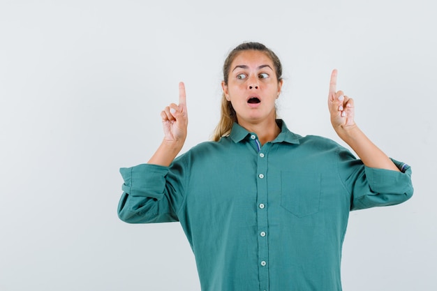 Foto gratuita mujer joven en blusa verde apuntando hacia arriba con los dedos índices y mirando sorprendido
