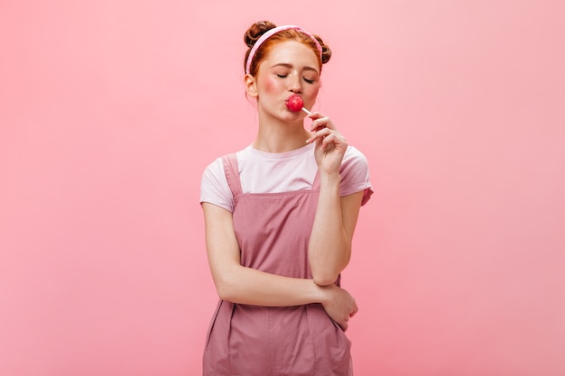 Mujer joven con bollos vestida con vestido rosa come piruleta sobre fondo aislado.