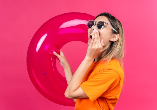 Foto gratuita una mujer joven y bonita alegre con una camiseta naranja con gafas de sol llamando a alguien mientras sostiene un anillo inflable rosa en una pared rosa