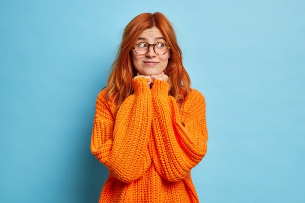 Una mujer joven y bonita con cabello rojo natural mantiene las manos debajo de la barbilla y mira a un lado, piensa en algo agradable, usa lentes ópticos y un jersey naranja de punto.