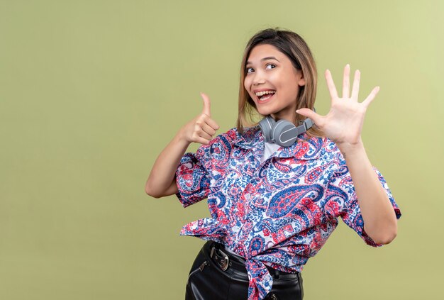 Una mujer joven y bonita en camisa estampada de paisley usando audífonos sonriendo y mostrando el número seis en una pared verde