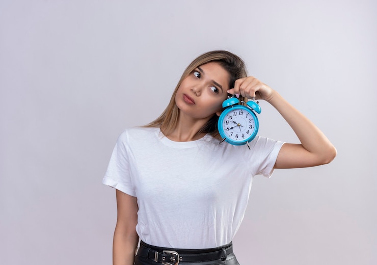Foto gratuito una mujer joven y bonita en camiseta blanca escuchando el tictac del reloj mientras sostiene el despertador azul en una pared blanca