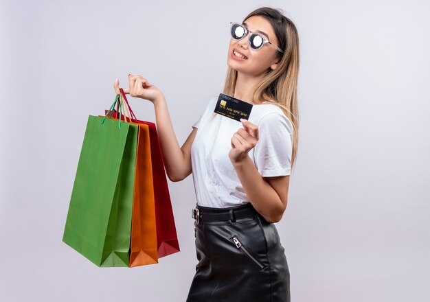 Una mujer joven y bonita en camiseta blanca con gafas de sol que muestra la tarjeta de crédito mientras sostiene bolsas de la compra en una pared blanca