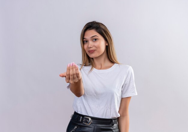 Una mujer joven y bonita en camiseta blanca llamando más cerca con gesto de la mano sobre una pared blanca