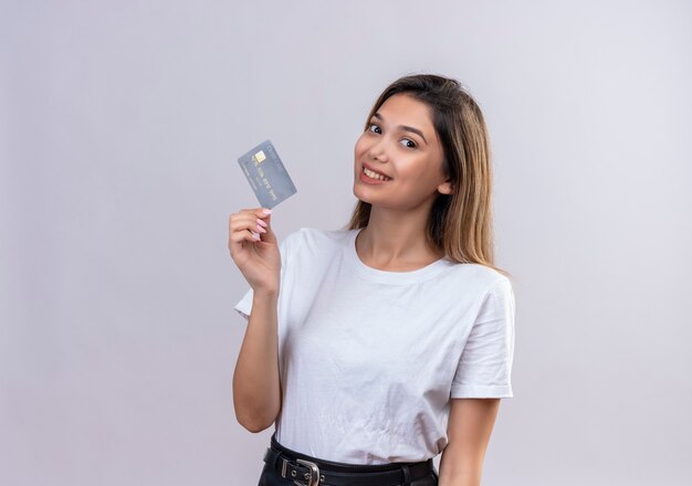 Una mujer joven y bonita en camiseta blanca que muestra la tarjeta de crédito en una pared blanca