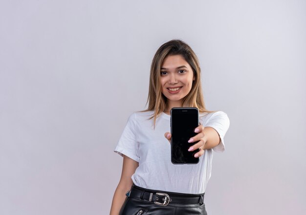 Una mujer joven y bonita en camiseta blanca sonriendo mientras muestra un espacio en blanco del teléfono móvil en una pared blanca