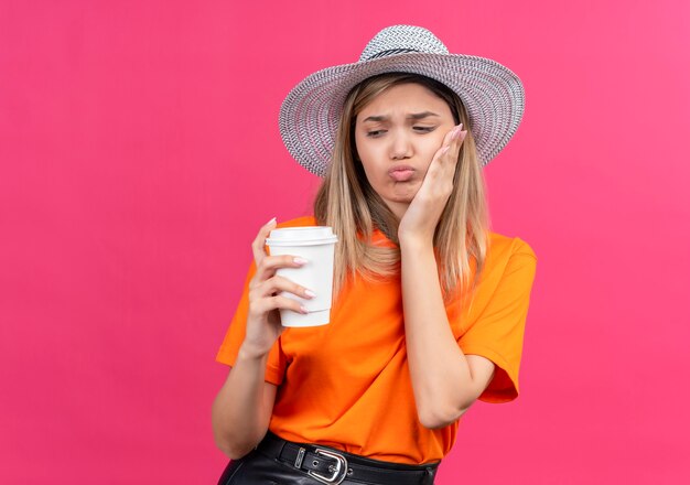 Una mujer joven y bonita en una camiseta naranja con sombrero para el sol manteniendo la mano sobre los dientes mientras sostiene un vaso de plástico en una pared rosa