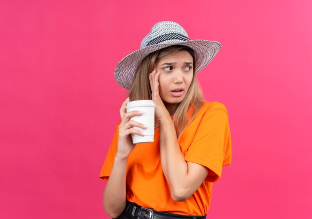 Una mujer joven y bonita confundida en una camiseta naranja con sombrero para el sol mirando de lado mientras sostiene un vaso de plástico en una pared rosa