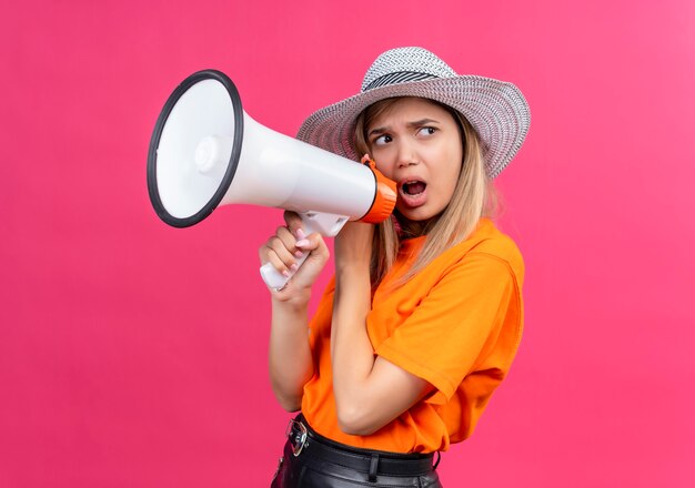 Una mujer joven y bonita enfadada en una camiseta naranja con sombrero para el sol hablando por megáfono