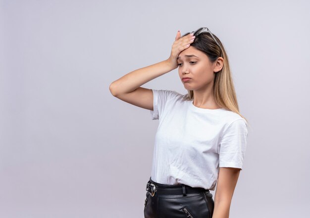 Una mujer joven y bonita estresante en camiseta blanca con gafas de sol en la cabeza manteniendo la mano en la cabeza sobre una pared blanca