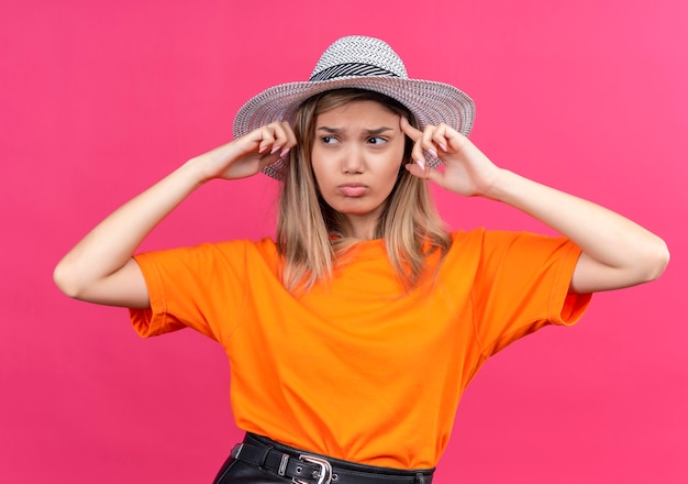 Una mujer joven y bonita estresante en una camiseta naranja con sombrero para el sol apuntando a su cabeza mientras mira de lado en una pared rosa