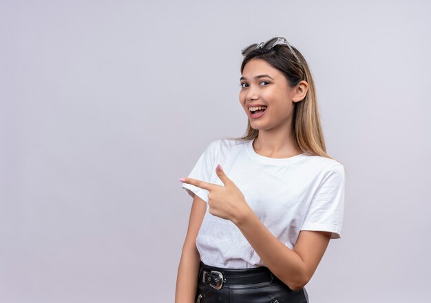 Una mujer joven y bonita feliz en camiseta blanca con gafas de sol en la cabeza apuntando a algo con el dedo índice en felicidad en una pared blanca