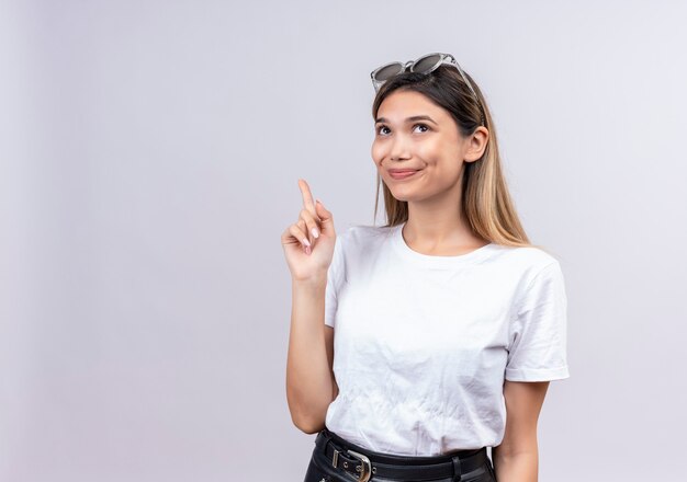 Una mujer joven y bonita feliz en camiseta blanca con gafas de sol en la cabeza levantando el dedo índice mientras tiene una sugerencia en una pared blanca