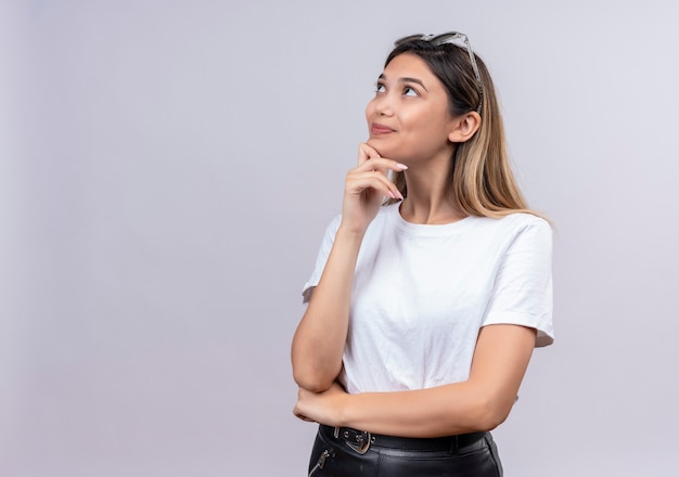 Una mujer joven y bonita feliz en camiseta blanca con gafas de sol en la cabeza manteniendo la mano en la barbilla mientras piensa en una pared blanca