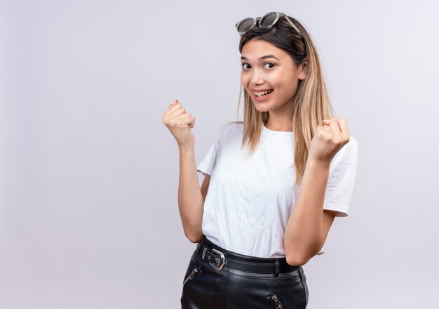 Una mujer joven y bonita feliz en camiseta blanca con gafas de sol en la cabeza mostrando los puños cerrados mientras mira en una pared blanca