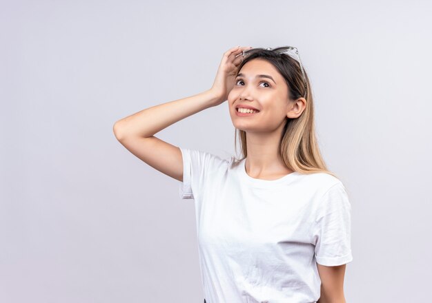 Una mujer joven y bonita feliz en camiseta blanca con gafas de sol en la cabeza pensando y mirando hacia arriba en una pared blanca