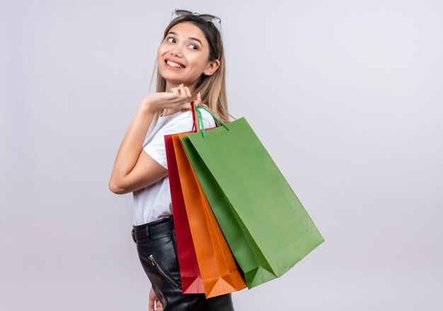 Una mujer joven y bonita feliz en camiseta blanca con gafas de sol en la cabeza sosteniendo bolsas de la compra en una pared blanca