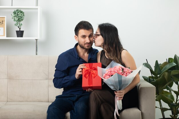 Mujer joven y bonita con gafas ópticas sosteniendo un ramo de flores y besando a un hombre guapo con una caja de regalo sentada en el sofá de la sala de estar en el día internacional de la mujer de marzo