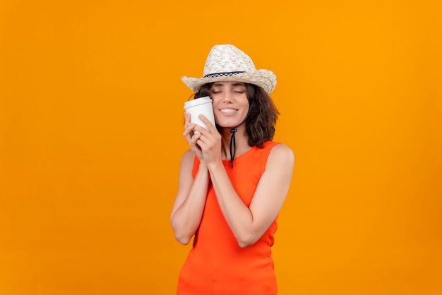 Una mujer joven y bonita con el pelo corto en una camisa naranja con sombrero para el sol abrazando una taza de café de plástico